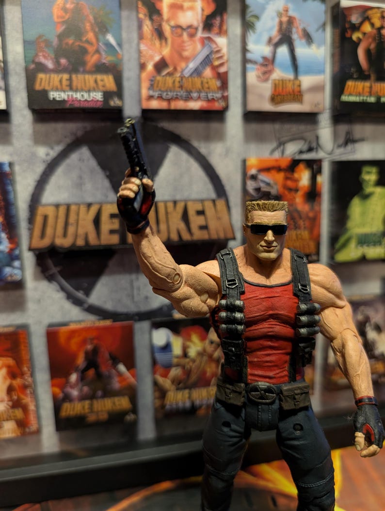 May include: A Duke Nukem action figure in a red tank top and black pants, holding a gun. The figure is in front of a Duke Nukem logo and several game box covers.