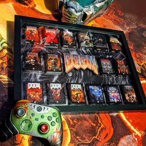 Op de afbeelding: Een ingelijste display met meerdere DOOM game cases, met een groot DOOM logo in het midden. Een aangepaste groene en rode Xbox controller staat op de voorgrond. De achtergrond heeft een oranje en zwart thema.