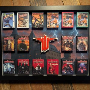 May include: A framed display featuring various Wolfenstein game boxes. The boxes are arranged against a backdrop of a castle and lightning. A large, red and white Wolfenstein logo is centered. The frame is black.