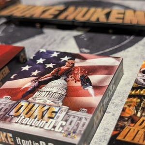 May include: A close-up of a game box featuring a muscular character with a gun, set against a backdrop of the U.S. Capitol and an American flag. The box reads "DUKE it out in D.C."
