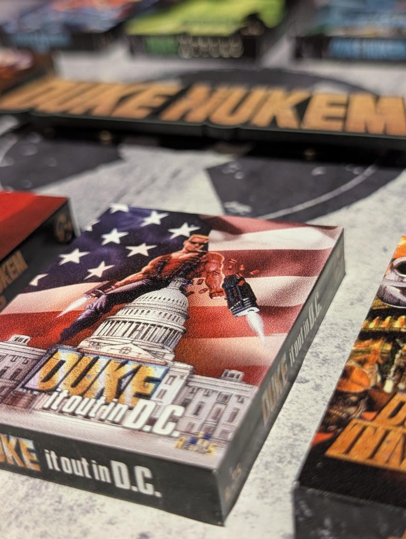 May include: A close-up of a game box featuring a muscular character with a gun, set against a backdrop of the U.S. Capitol and an American flag. The box reads "DUKE it out in D.C."