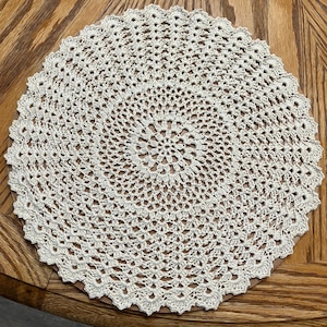 May include: A cream-colored, circular doily with a detailed crochet pattern. The doily features a central floral design and a scalloped edge. The handmade piece is placed on a wooden surface.