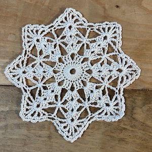 May include: A white crocheted doily with a starburst design. The doily is made with a delicate, intricate pattern and has a scalloped edge.
