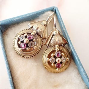 Peut inclure: Une paire de boucles d'oreilles antiques dorées avec un motif circulaire. Chaque boucle d'oreille présente un groupe central de pierres précieuses roses et claires, entouré d'une bordure décorative. Les boucles d'oreilles sont présentées dans une boîte bleue.