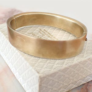 May include: A wide, oval-shaped gold-toned metal bangle bracelet with a smooth, matte finish. The bracelet features a hinged opening mechanism on one side and a secure clasp closure, presenting a clean, solid band design without any additional ornamentation.