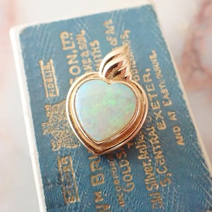 May include: A heart-shaped pendant with a gold frame and a pearly opal centre. The pendant has a small loop at the top for a chain. The opal has a subtle play of colour. The pendant is on a blue background with gold text.