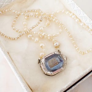 May include: A vintage pearl necklace with a rectangular pendant. The pendant has a silver frame and a blue background with a gold design. The necklace is displayed in a white box.