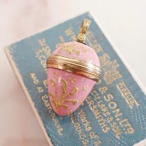 以下が含まれることがあります： ピンクのエナメル仕上げに金色の花柄が描かれ、中央に金色の金属バンドがあしらわれた卵型のペンダント。ペンダントの上部には、チェーンに取り付けるための小さな装飾的な金属製のバチカンが付いています。