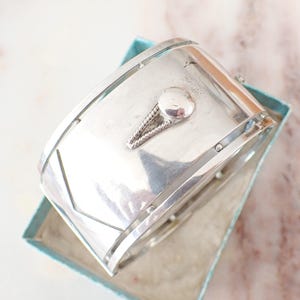 Può includere: Un ampio bracciale a fascia in argento lucido caratterizzato da una superficie liscia e curva con un accento triangolare centrale composto da filo intrecciato e un piccolo perno circolare. Il bracciale presenta linee geometriche.