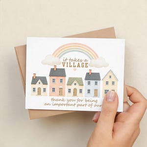 Puede incluir: Una tarjeta de felicitación con una ilustración en acuarela de cinco casas coloridas bajo un arcoíris y el texto "it takes a VILLAGE". La tarjeta se sostiene en una mano y está dentro de un sobre marrón. El texto "thank you for being an important part of our" también está en la tarjeta.