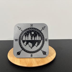 May include: A square, gray coaster with a compass design. The design features a mountain range, trees, and a full moon, with arrows pointing North, South, East, and West. The coaster sits on a round, wooden coaster.