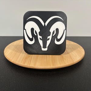 May include: A black square with a white ram head logo, resting on a round, light brown wooden coaster. The ram head features curved horns and a detailed face. The coaster sits on a black surface.