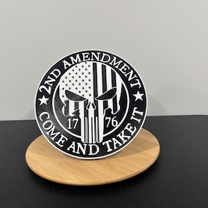 May include: A black and white circular sticker featuring a skull design with an American flag motif. The text "2nd Amendment Come and Take It" is displayed around the skull. The sticker is on a wooden coaster.