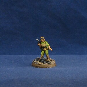 May include: Miniature figure of a person in green clothing, holding a weapon. The figure stands on a small, round base with a textured surface. The figure is painted with detailed features and is set against a blue background.