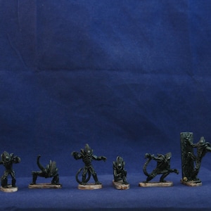 May include: A collection of seven dark green alien figurines in various poses, each mounted on a small, rectangular base. The figures are set against a deep blue backdrop, showcasing their detailed designs.