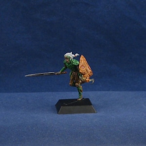 May include: Miniature figure of a fantasy warrior in green armor, holding a sword and shield. The figure has white hair and is posed in a dynamic stance on a black base. The shield is orange with a detailed design.