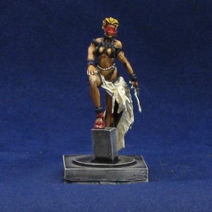 May include: A miniature figurine of a warrior woman with a blindfold, standing on a pedestal. She wears a dark outfit with a white cloth draped over her arm and holds a sword. The figure is painted with detailed shading.