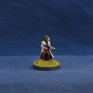 May include: Miniature figure of a person in a white coat and maroon dress, holding a futuristic weapon. The figure stands on a circular base covered in green flocking, against a blue backdrop. The miniature is likely for tabletop gaming.