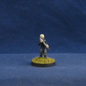 May include: Miniature figurine of a humanoid figure with a pale head, wearing a black jacket and gray pants. The figure holds a silver and brown cylindrical object. It stands on a circular base covered in green and yellow vegetation.