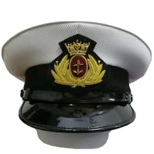 Pode incluir: Chapéu branco de oficial da marinha com viseira e faixa pretas. O chapéu apresenta um emblema dourado com uma coroa, uma âncora e folhas de louro. O chapéu tem um acabamento brilhante e um design estruturado.