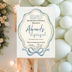Op de afbeelding: Een welkomstbord op een ezel met een aquarelontwerp. Het bord heeft tekst in het Engels en Arabisch, met een blauw en groen kleurenschema. Op het bord staat "Welkom bij Ahmed's Ageegah".