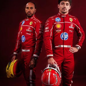 May include: Two Formula 1 drivers in red racing suits with white accents and sponsor logos. One holds a yellow and red helmet, the other a red helmet. The suits feature the IBM, HP, and UniCredit logos. The background is a deep red.