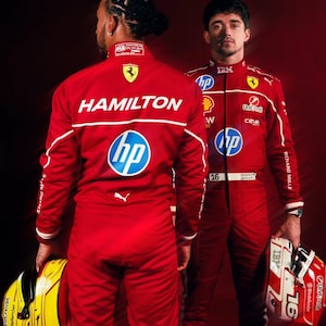 May include: Two Formula 1 racing drivers in red racing suits with white accents. One suit has "HAMILTON" printed on the back, the other has "CHARLES LECLERC" printed on the front. Both suits feature sponsor logos and the number "16". One driver holds a yellow helmet, the other a red and white helmet.