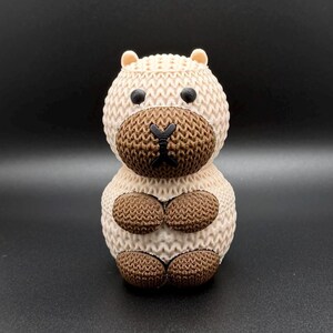 May include: A knitted capybara figurine in shades of beige and brown. The capybara has a textured body, a dark brown face, and small, rounded ears. Its front paws are folded, and it has a cute, friendly appearance.