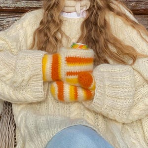 Pode incluir: Uma pessoa vestindo um suéter de malha creme e luvas combinando. As luvas são listradas com laranja e amarelo. A pessoa está segurando uma lata de bebida.
