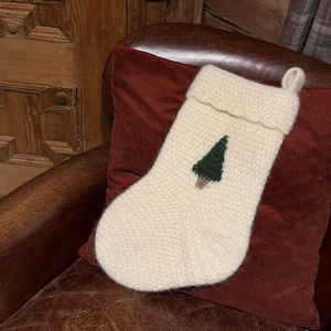 Könnte beinhalten: Cremefarbener gestrickter Weihnachtsstrumpf mit einem grün-braunen gestickten Baumdesign. Der Strumpf wird auf einem weinroten Kissen und einem braunen Ledersessel präsentiert, was ein gemütliches Urlaubsthema suggeriert.