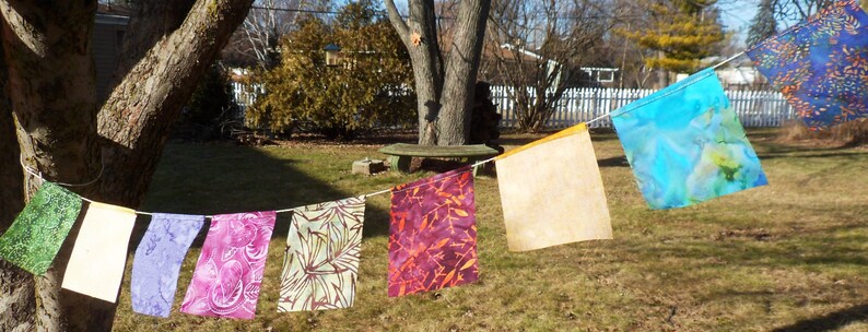 May include: A colorful banner of square fabric flags strung on a white cord. The flags display various patterns and colors, including green, purple, pink, and blue, with leaf and abstract designs. The banner is hanging outdoors in a sunny yard.