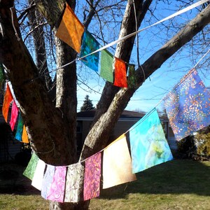May include: A tree decorated with colorful fabric flags. The flags are various colors, including orange, blue, green, and purple, and are strung on a white rope. The tree is in a yard with a house in the background.