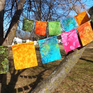 May include: A colorful banner of square fabric flags in various colors, including green, orange, blue, and pink, hangs from a string. The flags have patterned designs and are displayed outdoors, attached to a tree branch.