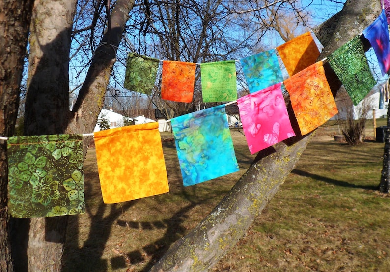 May include: A colorful banner of square fabric flags in various colors, including green, orange, blue, and pink, hangs from a string. The flags have patterned designs and are displayed outdoors, attached to a tree branch.