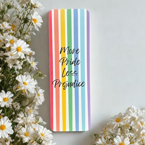 Puede incluir: Marcapáginas rectangular con diseño de rayas arcoíris y el texto "More Pride Less Prejudice" en escritura negra. Margaritas blancas enmarcan el marcapáginas sobre un fondo blanco.
