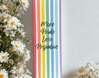 May include: A rainbow-striped bookmark with the words "More Pride Less Prejudice". The bookmark is flanked by white daisy flowers against a white backdrop.