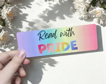 May include: A rectangular bookmark with a rainbow gradient. The text "Read with PRIDE" is printed on the bookmark in black and rainbow colours. The bookmark is held by a hand, with white flowers in the background.