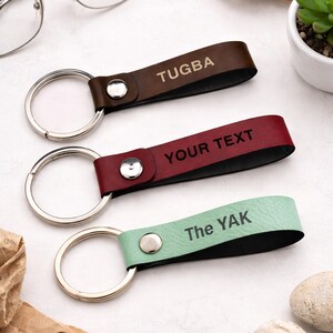 Op de afbeelding: Drie leren sleutelhangers in bruin, bordeauxrood en mintgroen, elk met een zilveren sleutelring. De bruine sleutelhanger leest "TUGBA", de bordeauxrode "YOUR TEXT" en de groene "The YAK".