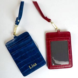 May include: Two leather ID badge holders with lanyards. One is navy blue with gold lettering that says "Lisa", and the other is red. Both have a crocodile texture and gold-coloured hardware.