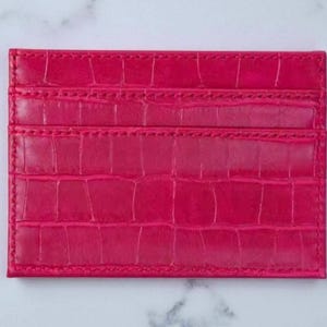 Puede incluir: Un tarjetero rosa brillante con textura de piel de cocodrilo. La cartera rectangular tiene tres ranuras para tarjetas y est&aacute; hecha de un material liso y brillante. Los bordes est&aacute;n cuidadosamente cosidos.