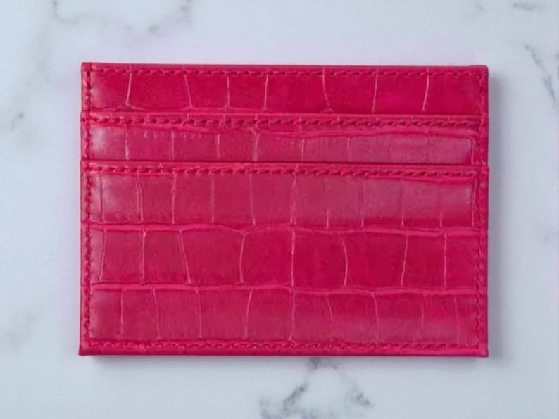 Puede incluir: Un tarjetero rosa brillante con textura de piel de cocodrilo. La cartera rectangular tiene tres ranuras para tarjetas y est&aacute; hecha de un material liso y brillante. Los bordes est&aacute;n cuidadosamente cosidos.