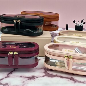 May include: A collection of oval cosmetic bags in shades of black, brown, burgundy, and pink. The bags have a clear, transparent body, a zip closure, and the name "Lisa" printed on top. Gold-tone hardware is used on the bags.