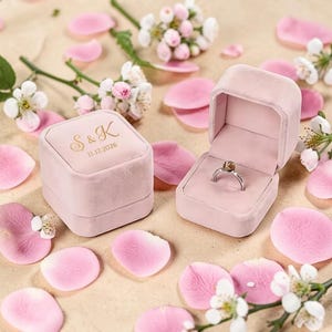 Könnte beinhalten: Eine rosa Samt-Ringbox mit den Initialen "S & K" und dem Datum "11.12.2026" in goldfarbenen Buchstaben. Die Box ist geöffnet und zeigt einen silbernen Ring mit einem Edelstein. Rosa Rosenblätter und weiße Blüten umgeben die Box.