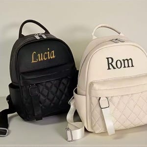 Peut inclure: Deux petits sacs à dos personnalisés. L'un est noir avec "Lucia" brodé en or, l'autre est crème avec "Rom" brodé en noir. Les deux ont un motif matelassé sur la poche avant et des bretelles réglables.