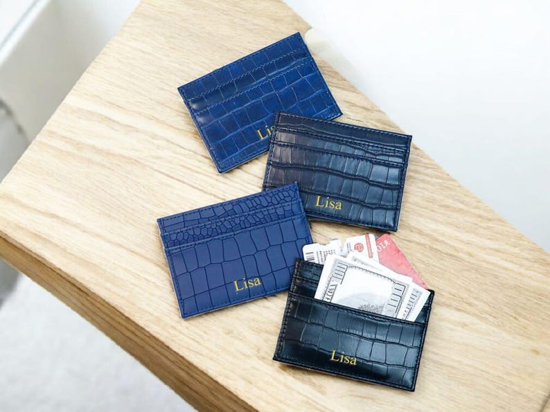 Puede incluir: Cuatro tarjeteros azules con textura de piel de cocodrilo se muestran sobre una superficie de madera. Cada tarjetero tiene el nombre "Lisa" impreso en dorado. Un tarjetero est&aacute; abierto, revelando tarjetas y efectivo.