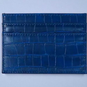 Puede incluir: Un tarjetero azul con textura de piel de cocodrilo. El tarjetero tiene m&uacute;ltiples ranuras para tarjetas y forma rectangular. El color es un azul profundo y vibrante.