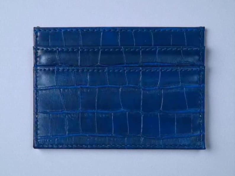 Puede incluir: Un tarjetero azul con textura de piel de cocodrilo. El tarjetero tiene m&uacute;ltiples ranuras para tarjetas y forma rectangular. El color es un azul profundo y vibrante.