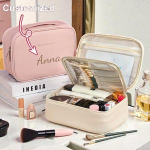 Op de afbeelding: Een roze gepersonaliseerde make-up tas met de naam "Anna" erop gedrukt. De tas is open en onthult diverse cosmetica en borstels. De tas heeft een handvat en een ritssluiting. De tekst "Beauty Preservation" staat op de tas.