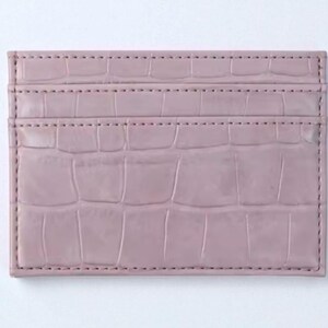 Puede incluir: Un tarjetero de color malva con textura de piel de cocodrilo. La cartera rectangular tiene tres ranuras para tarjetas y est&aacute; cosida en los bordes. El fondo es blanco.