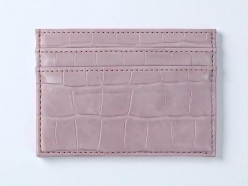 Puede incluir: Un tarjetero de color malva con textura de piel de cocodrilo. La cartera rectangular tiene tres ranuras para tarjetas y est&aacute; cosida en los bordes. El fondo es blanco.